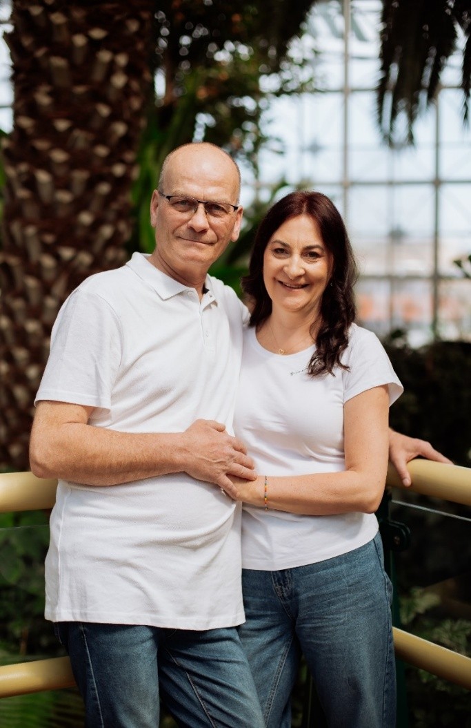 Kobieta o długich ciemnych włosach i mężczyzna w okuklarach, ubrani w białe T-shirty, stoją przytuleni
