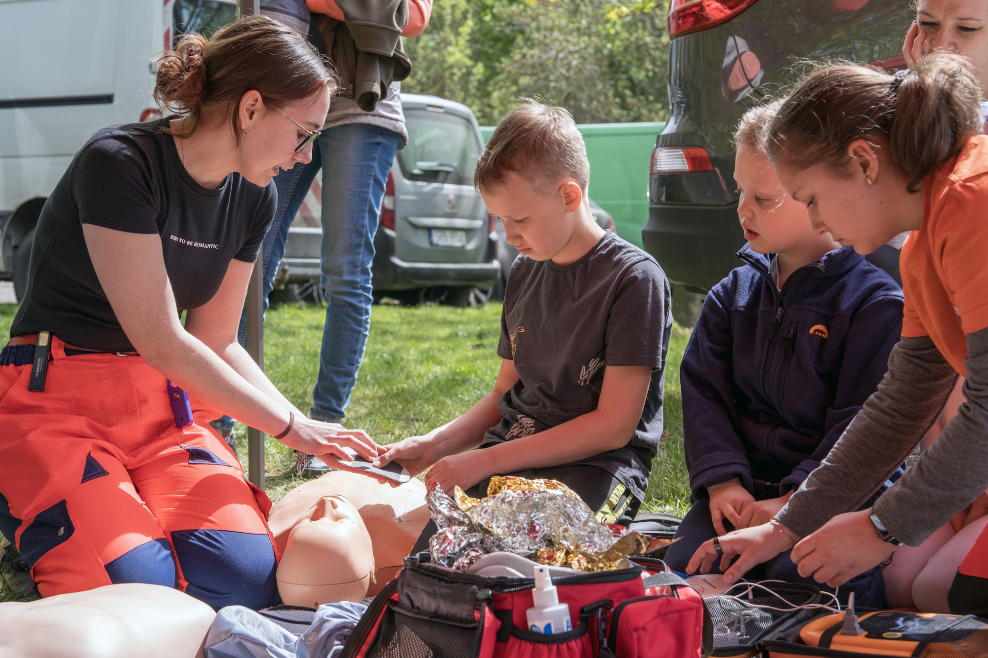troje dzieci klęczy i patrzy jak kobieta uczy pierwszej pomocy na fantomie