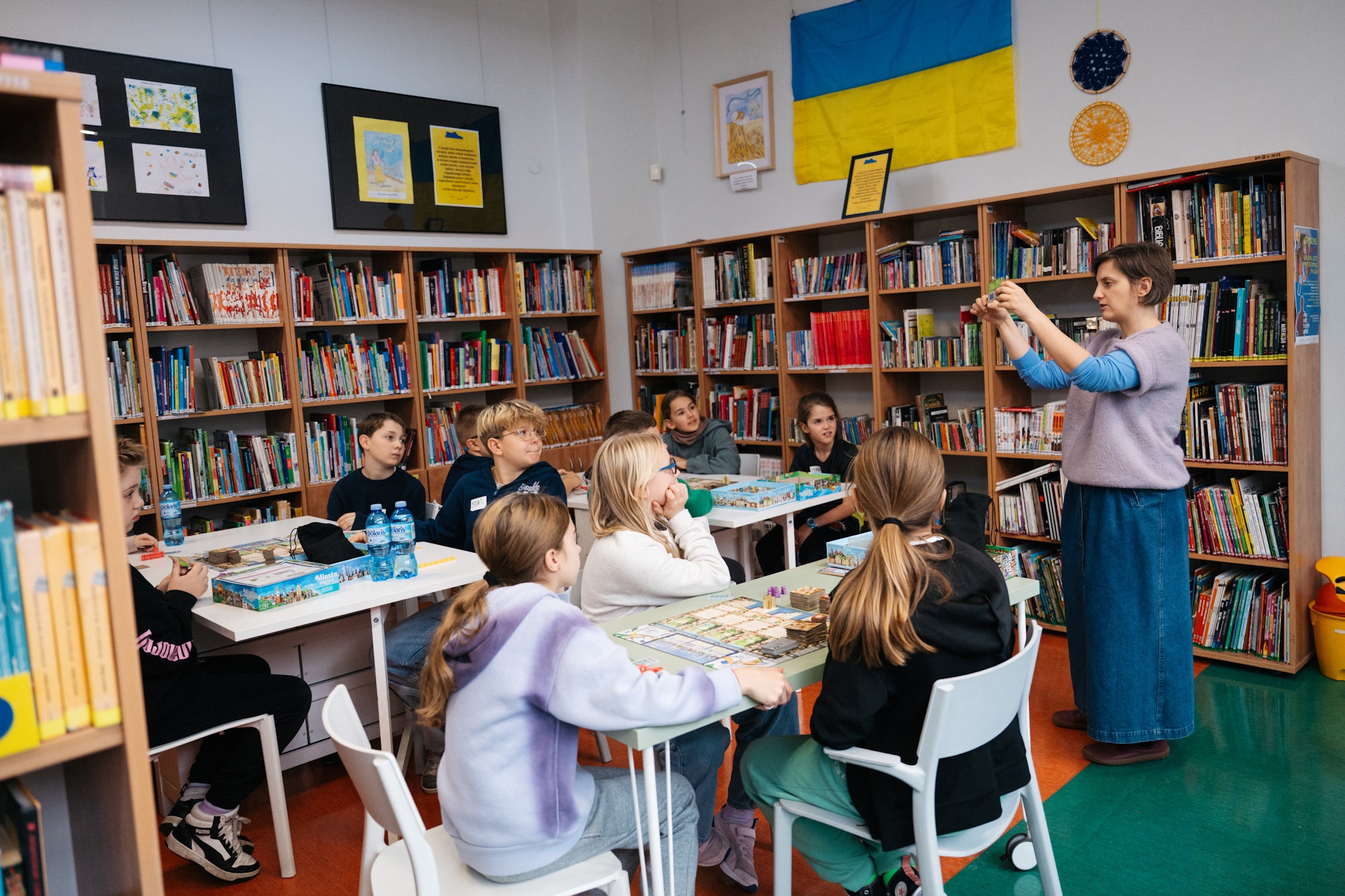 kobieta robi zdjęcie dzieciom siedzącym przy stolikach w bibliotece
