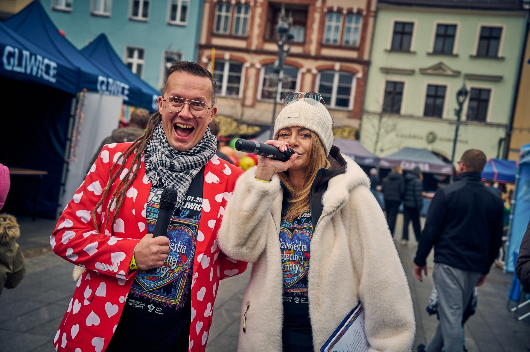 Uśmiechcięty mężczyzna w czerwonej marynarce z białymi serduszkami, obok niego kobieta z mikrofonem