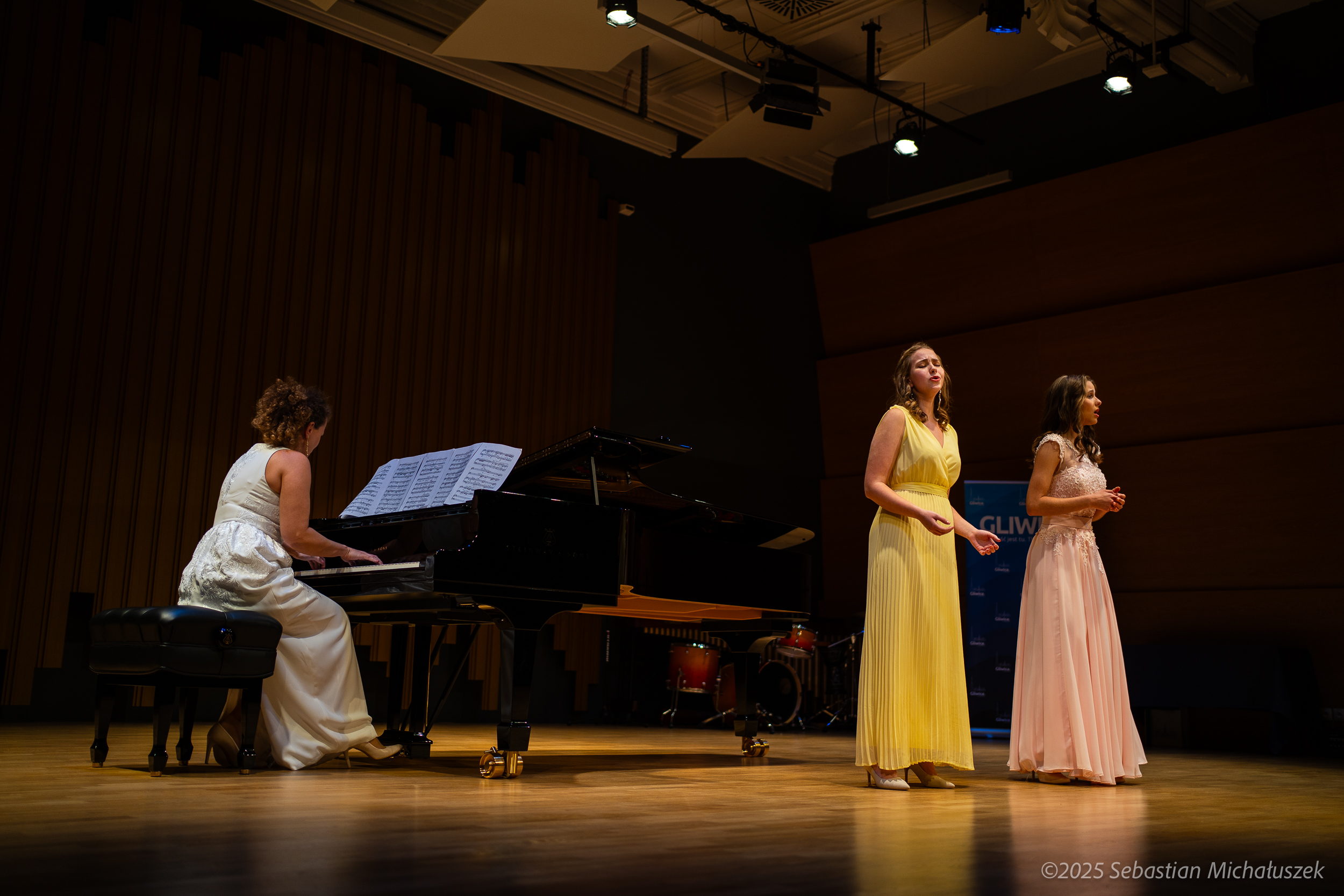 dwie solistki w długich sukniach: żółtej i różowej oraz kobieta przy pianinie