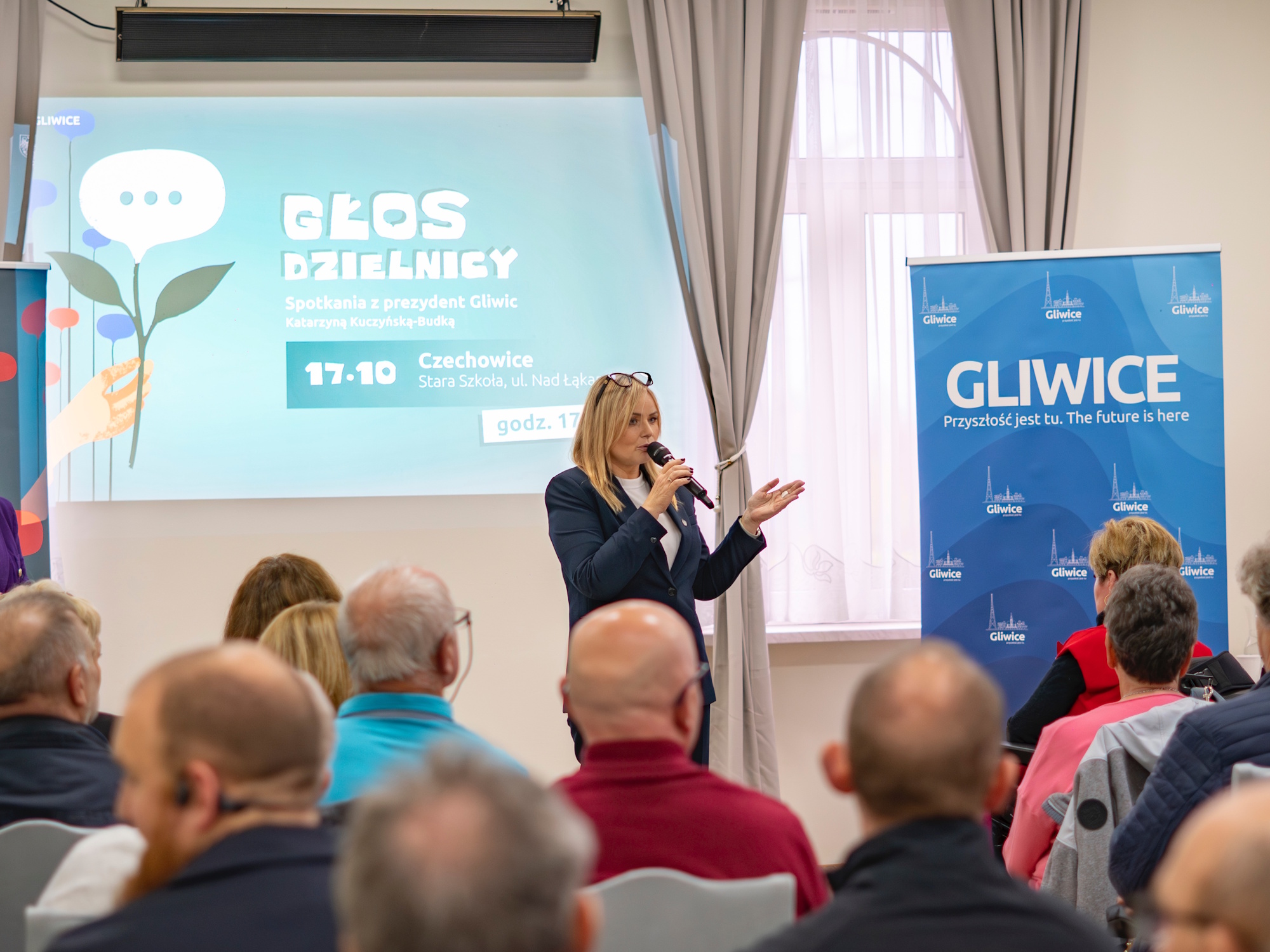 ludzie siedzą na krzesłach, prezydent stoi przed nimi i mówi do mikrofonu