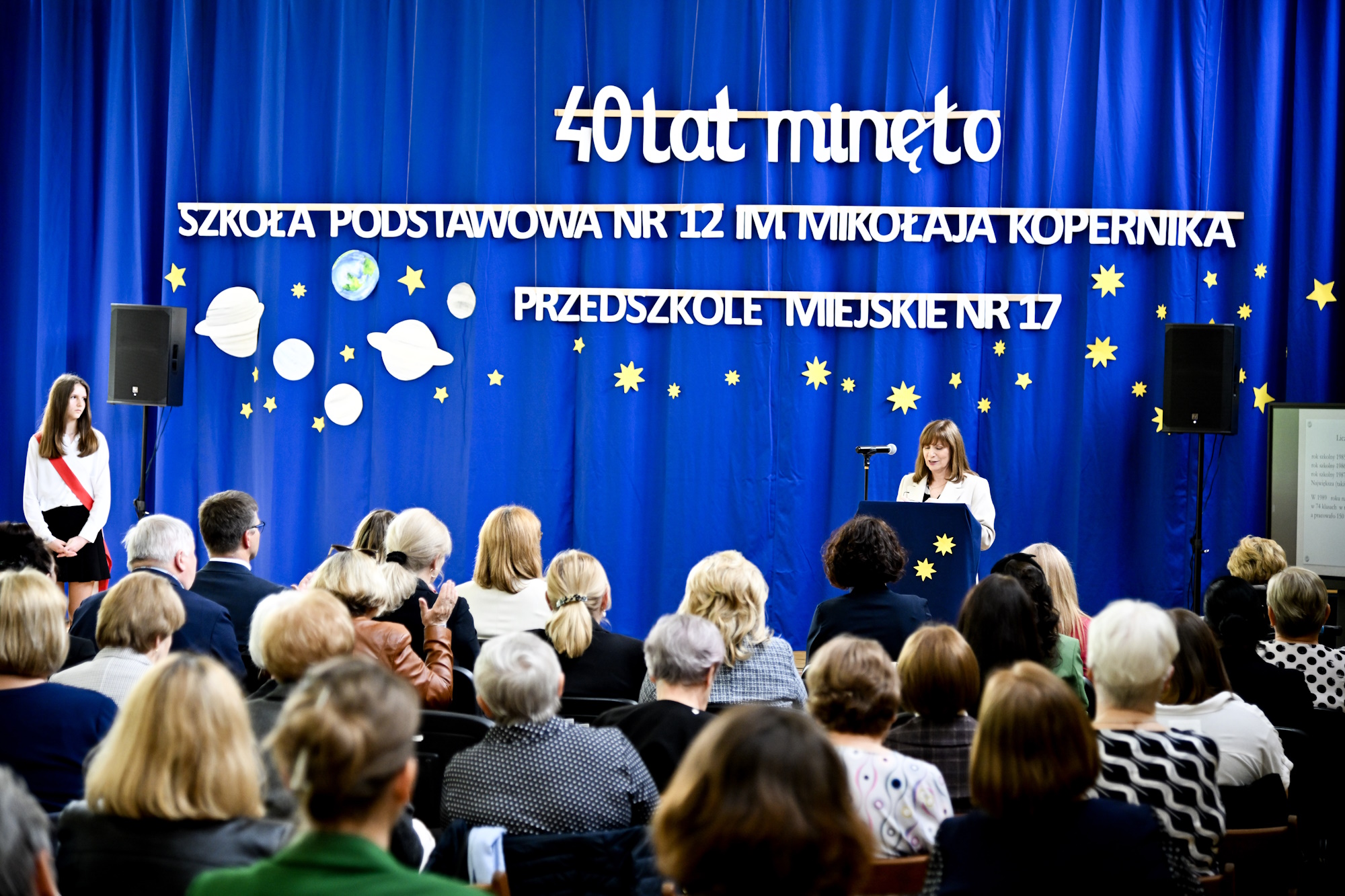 Ludzie w sali gimnastycznej szkoły podczas uroczystości, siedzą na krzesłach
